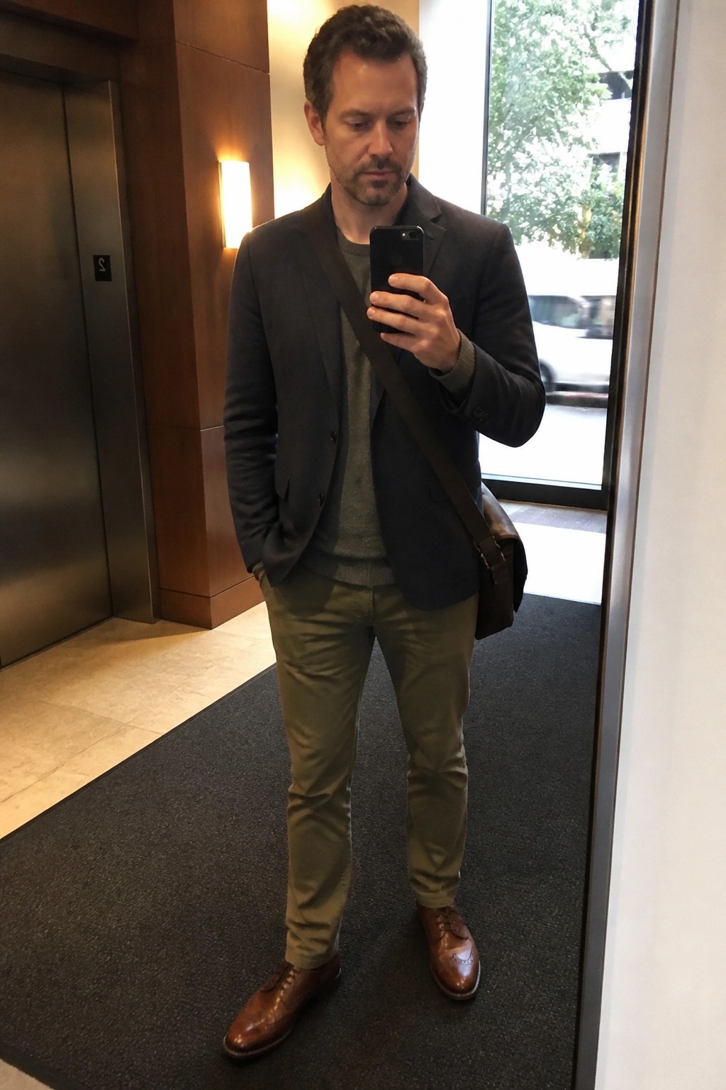 A man in a dark gray blazer over a gray sweater with olive pants and brown brogue shoes, carrying a brown messenger bag while holding a phone.