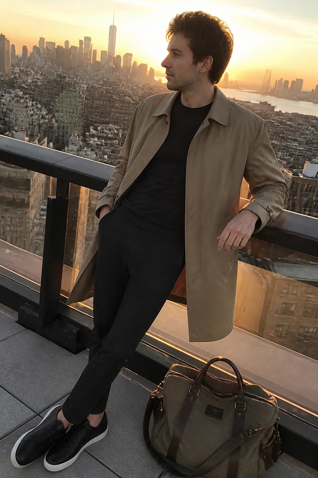 Man in tan coat over black attire on balcony overlooking NYC sunset with duffel bag
