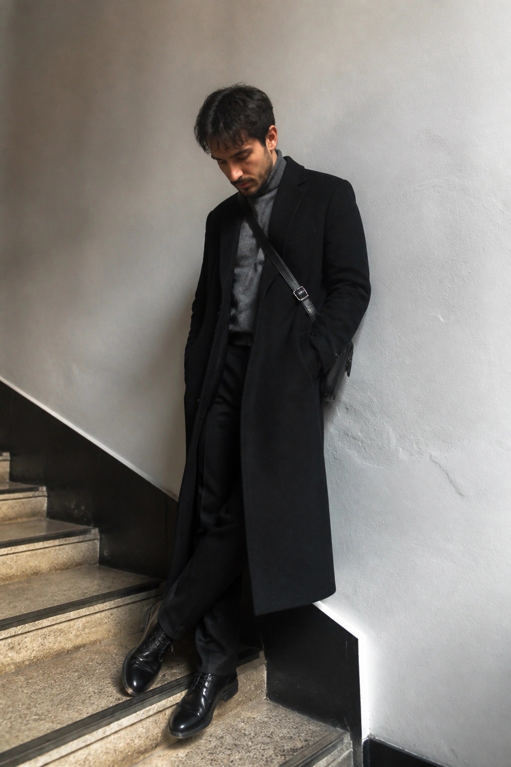 Man in black overcoat and gray turtleneck leaning on marble staircase wall