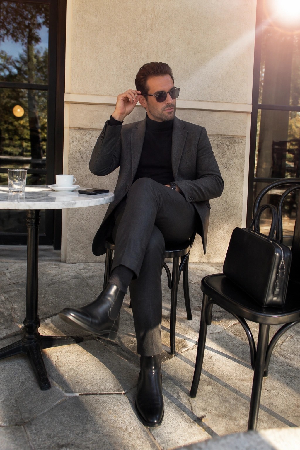 Man in gray suit and black turtleneck adjusts sunglasses at sunny cafe patio.