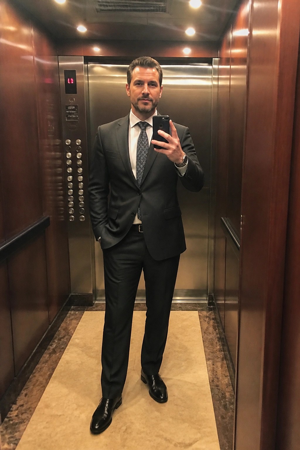 Man in dark suit taking selfie in wood-paneled elevator with metal doors.