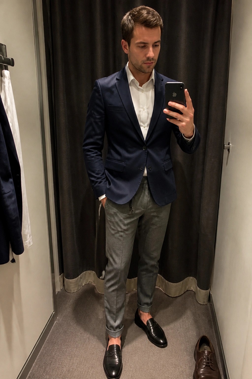 Man in navy blazer and gray pants taking mirror selfie in fitting room.