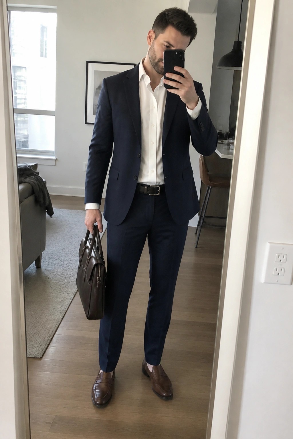 Man in navy suit holding brown briefcase and phone for mirror selfie in apartment