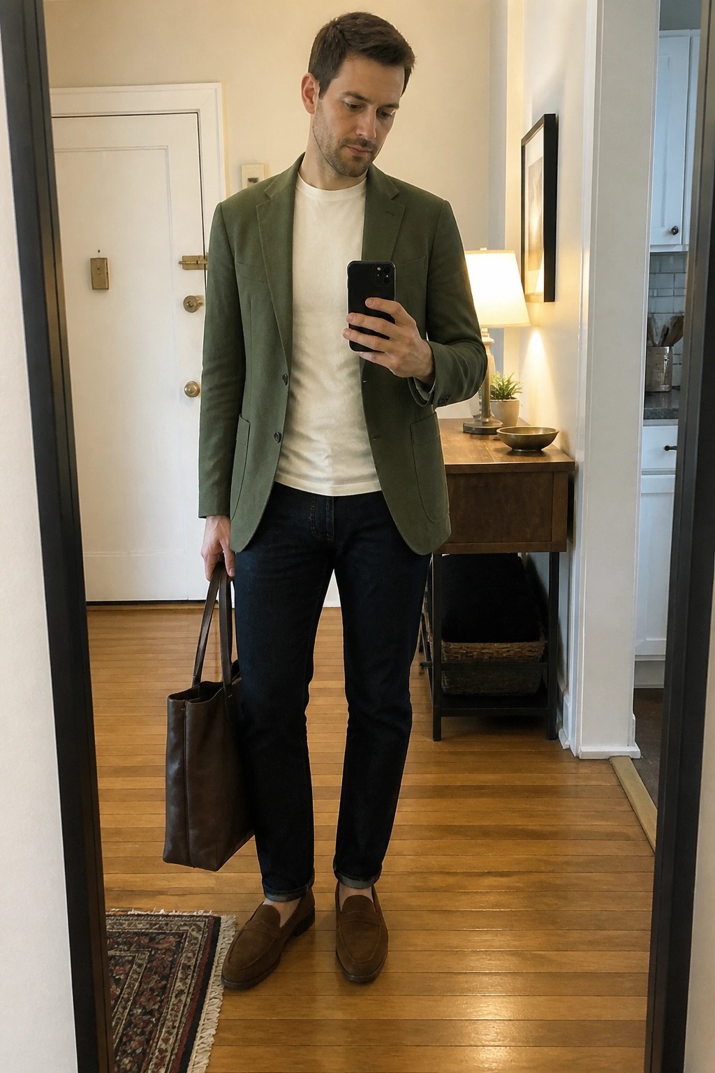 Man in green blazer and jeans holding phone and brown leather tote in hallway