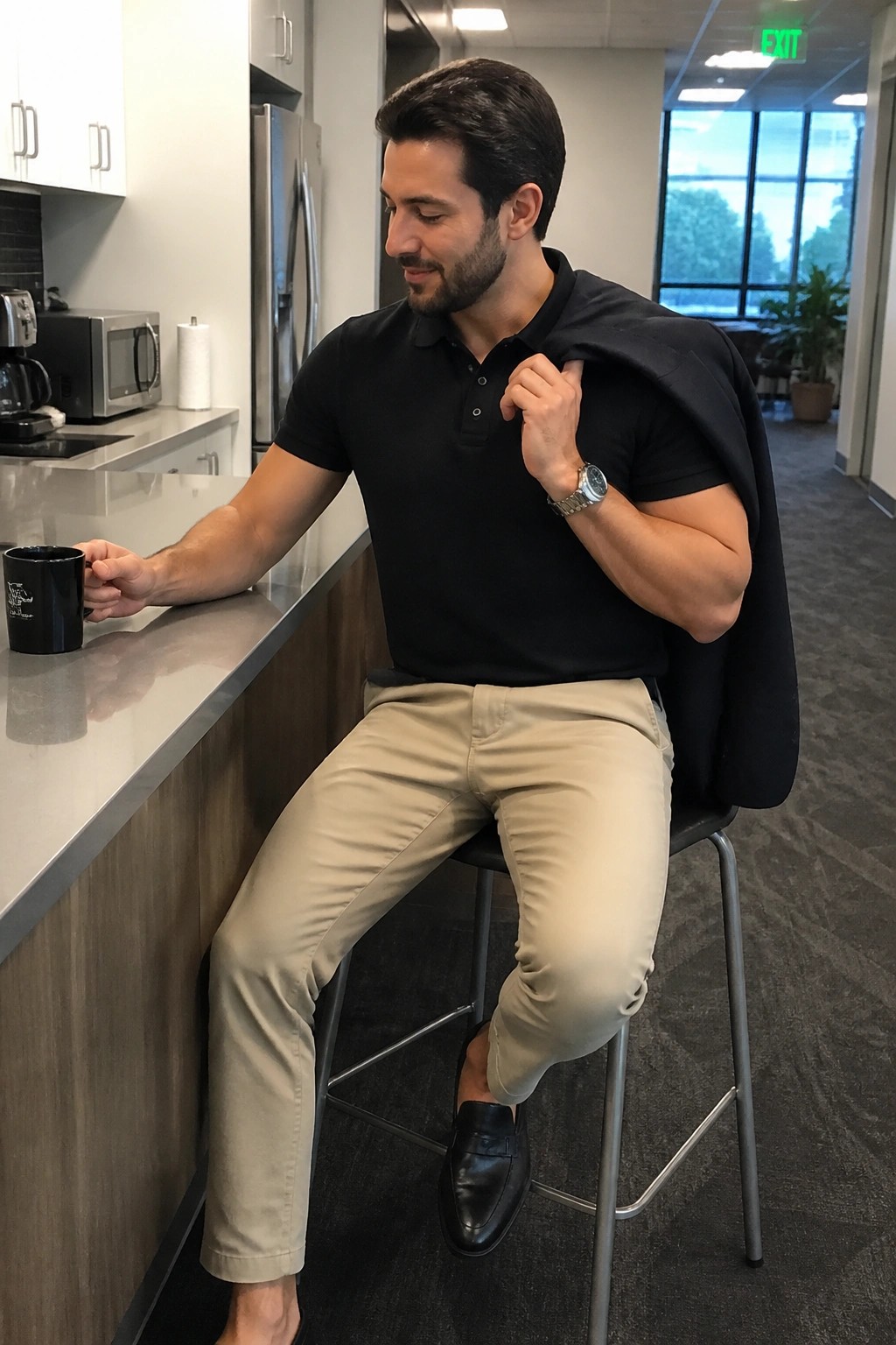Man in black polo shirt and khaki pants holds mug with jacket over shoulder in office kitchen