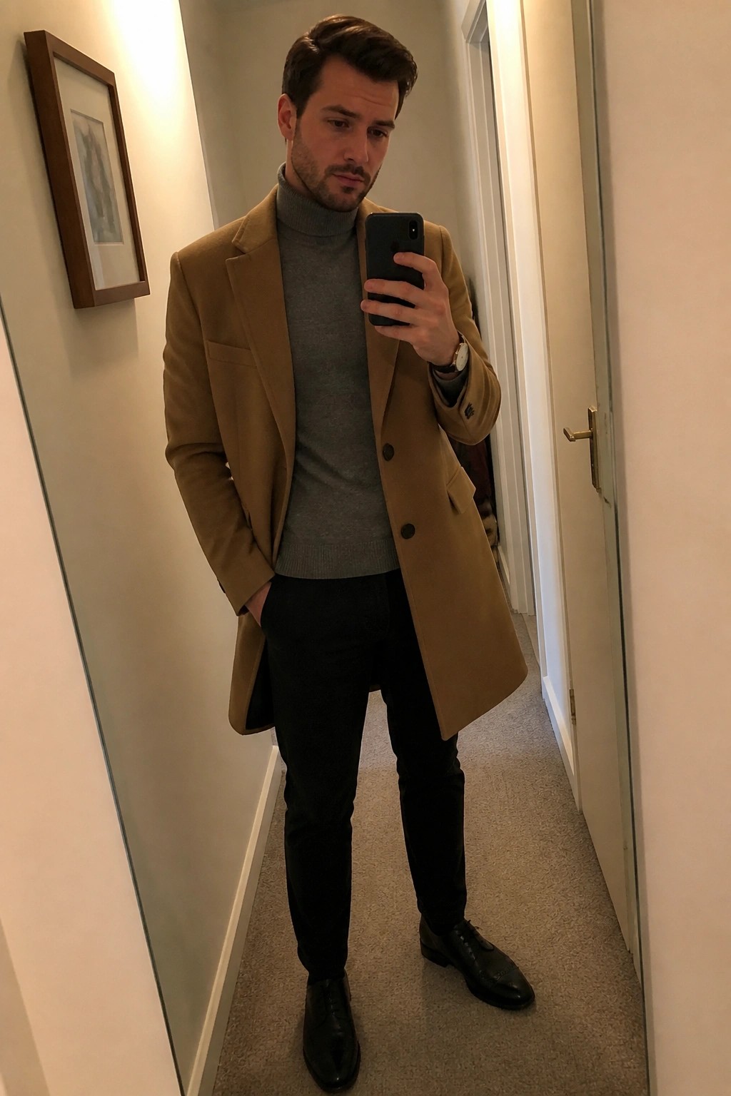 Man in tan overcoat and gray turtleneck taking mirror selfie in hallway