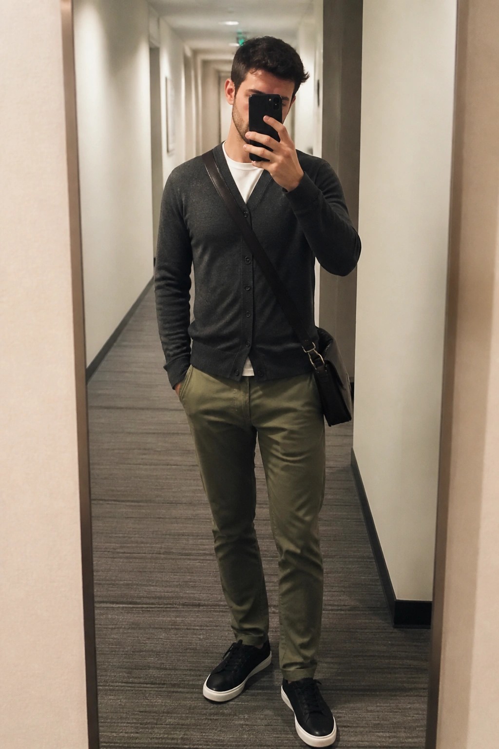 Man in gray cardigan and olive pants taking mirror selfie in hallway