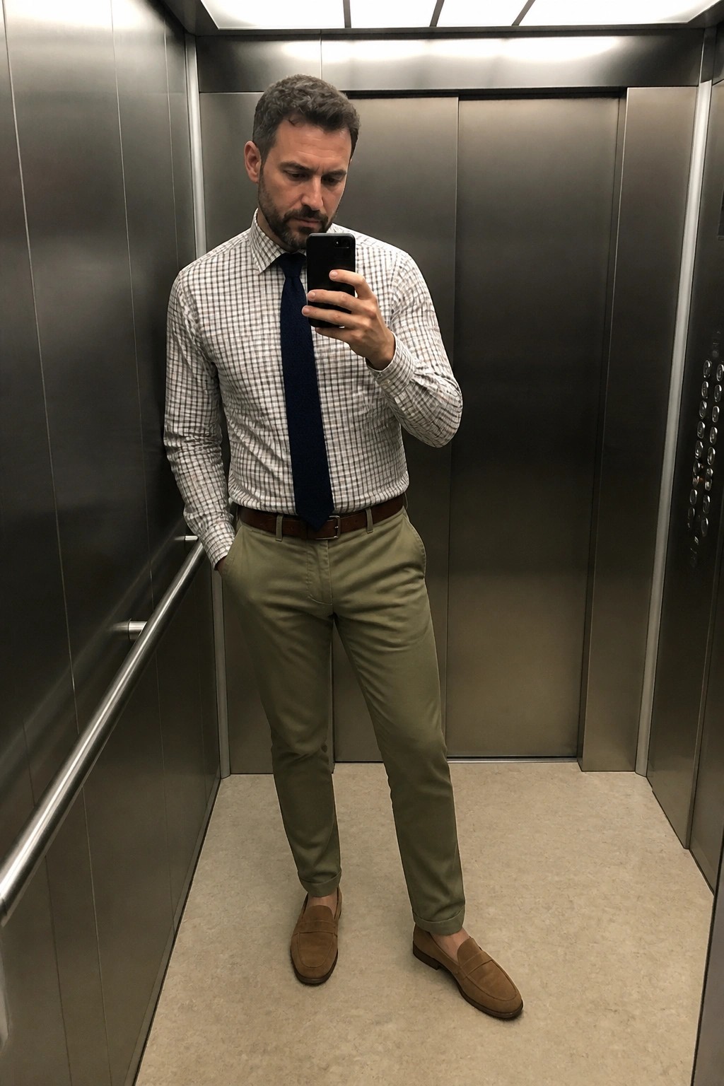 Man in checkered shirt and tie taking mirror selfie in elevator