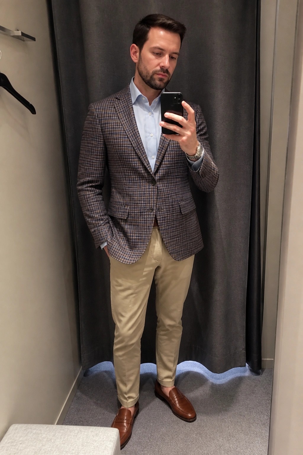 Man in plaid blazer and khaki pants taking mirror selfie in fitting room