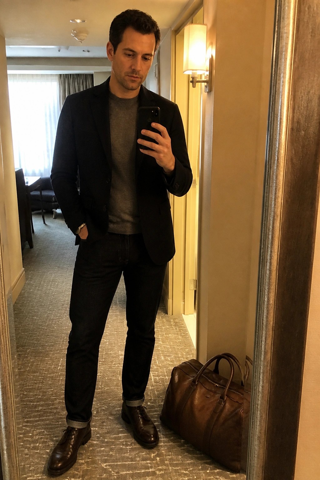Man in black blazer and gray sweater taking mirror selfie beside brown leather bag