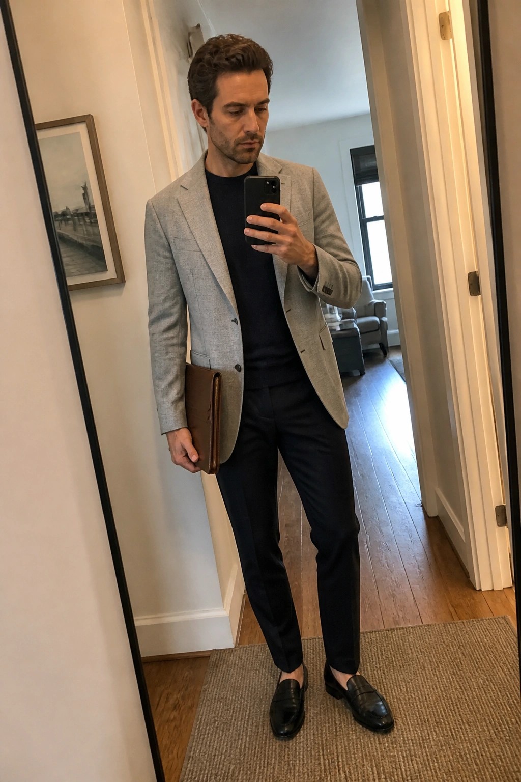 Man in gray blazer and navy outfit taking mirror selfie with brown leather portfolio.