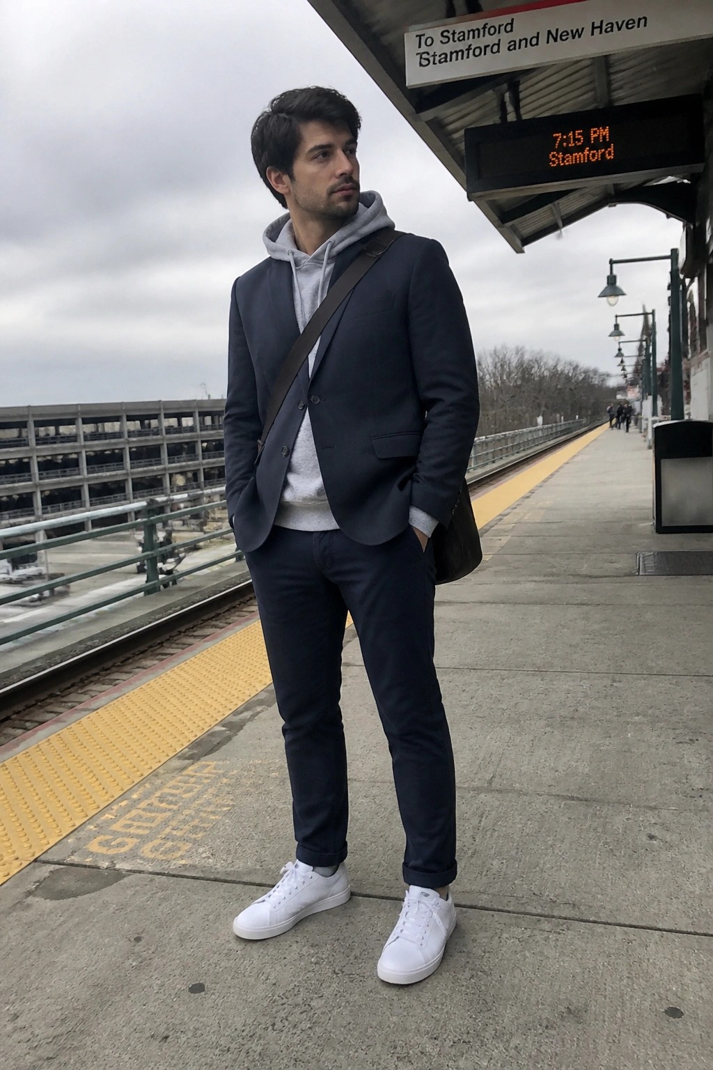 Man in navy blazer over grey hoodie with white sneakers on train platform
