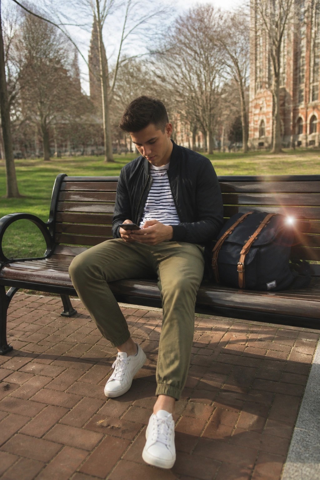 Young man in bomber jacket checks phone on park bench beside blue backpack