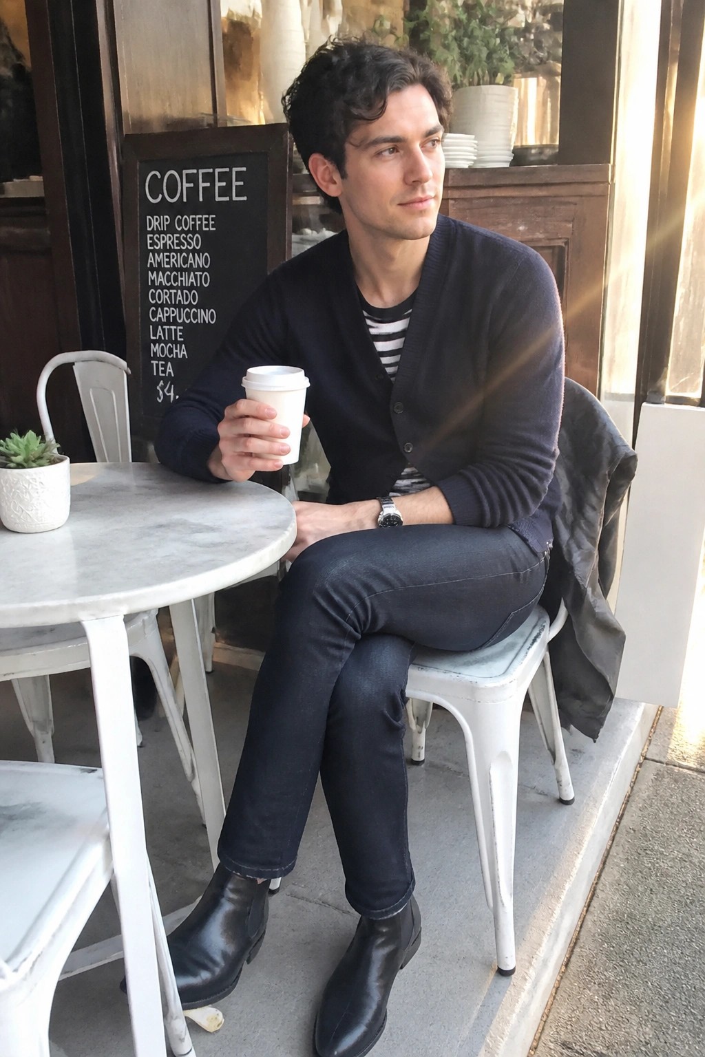 Man in navy cardigan holding white coffee cup at sunny outdoor cafe table