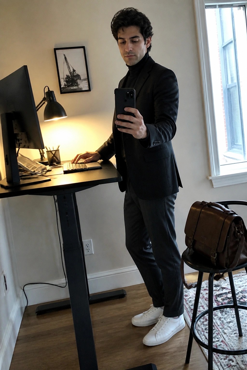 Man in black suit and white sneakers holding smartphone at standing desk with monitor