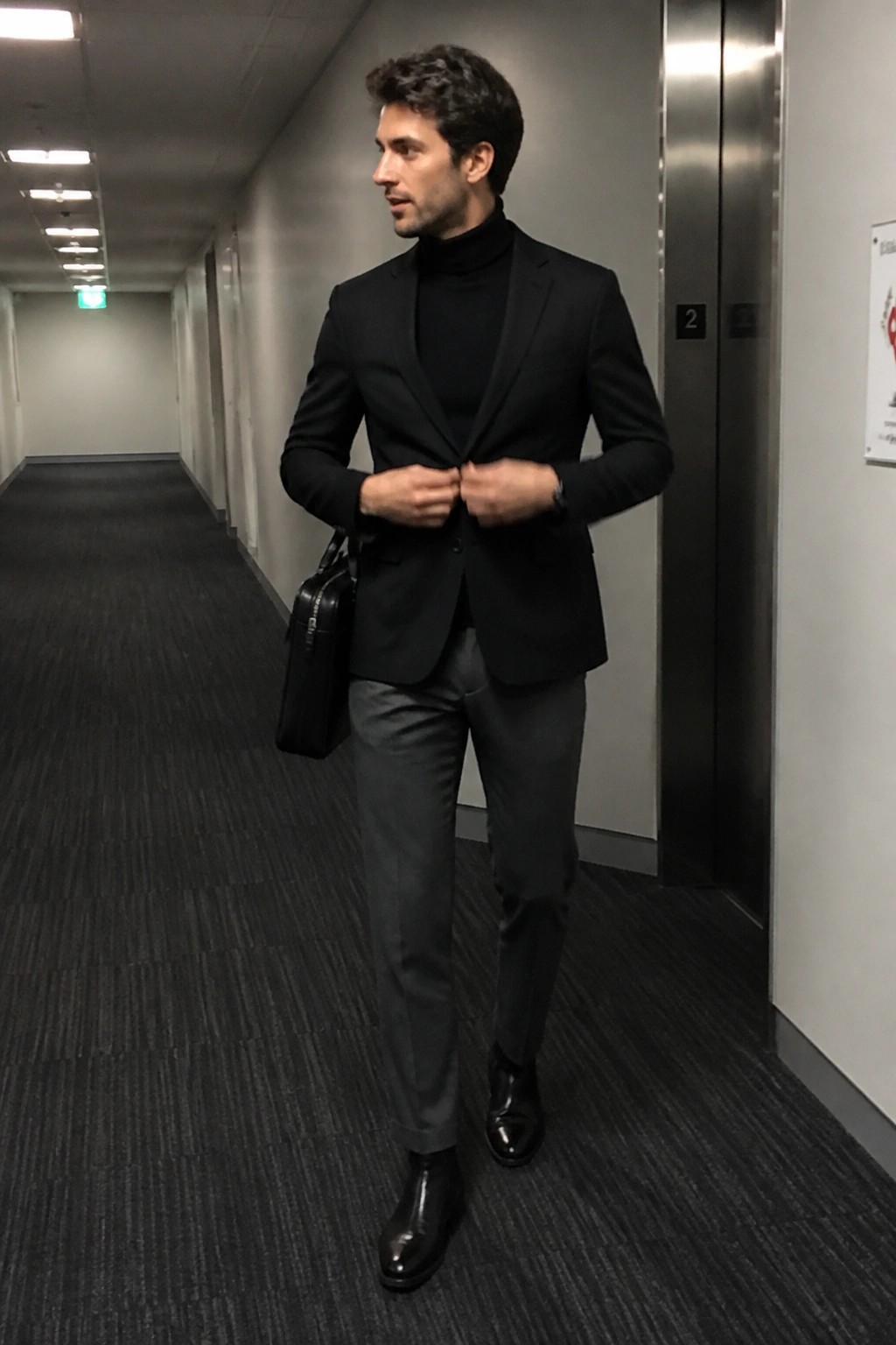 Man in black blazer over turtleneck adjusting jacket with briefcase in hallway