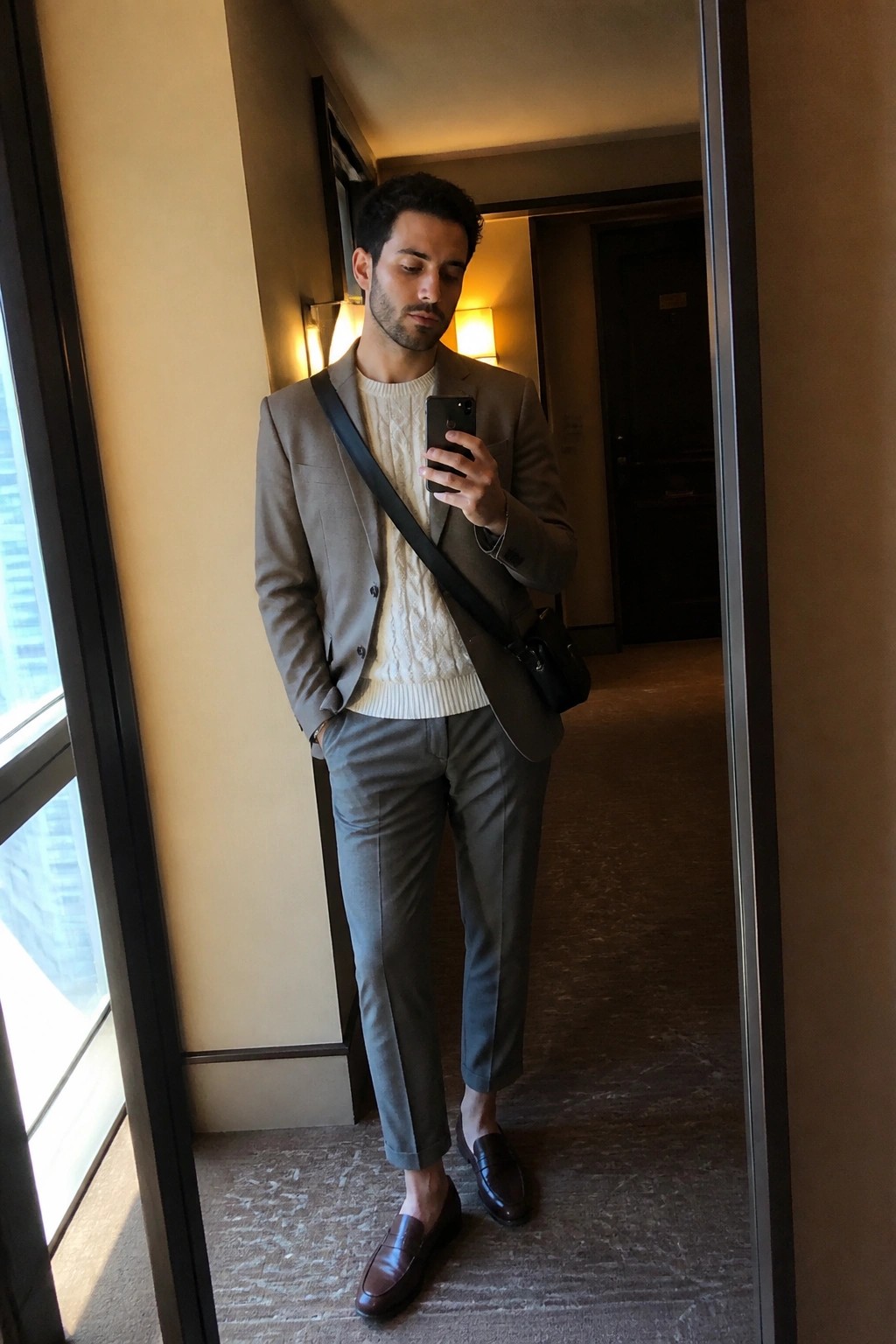 Man in gray blazer over white cable sweater taking mirror selfie in hallway