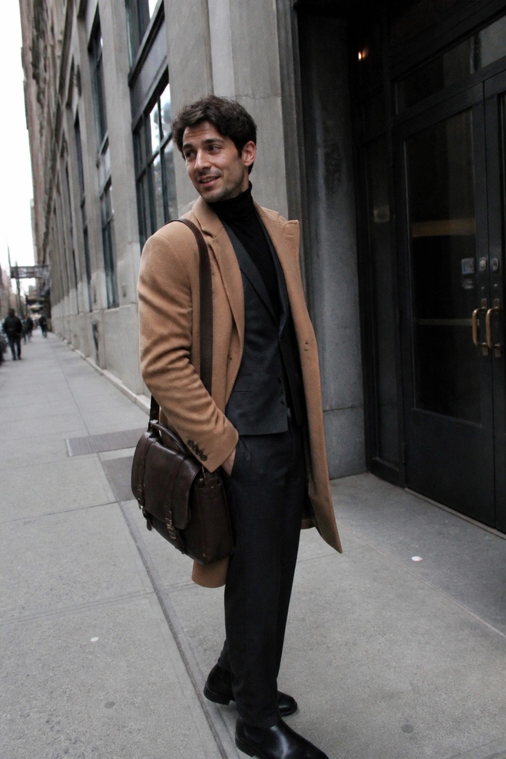 Man in tan overcoat and black suit with brown leather bag on sidewalk
