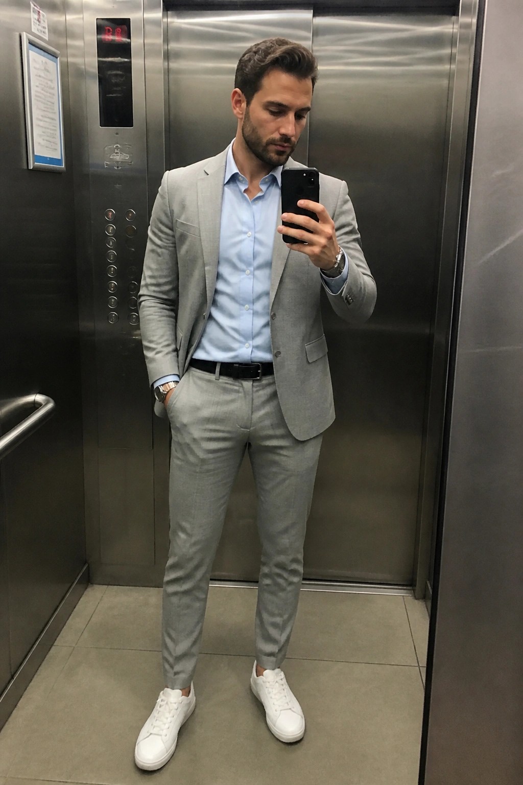 Man wearing a light gray tailored suit with matching trousers, pale blue button down shirt, black belt, and white sneakers standing in an elevator taking a mirror selfie.