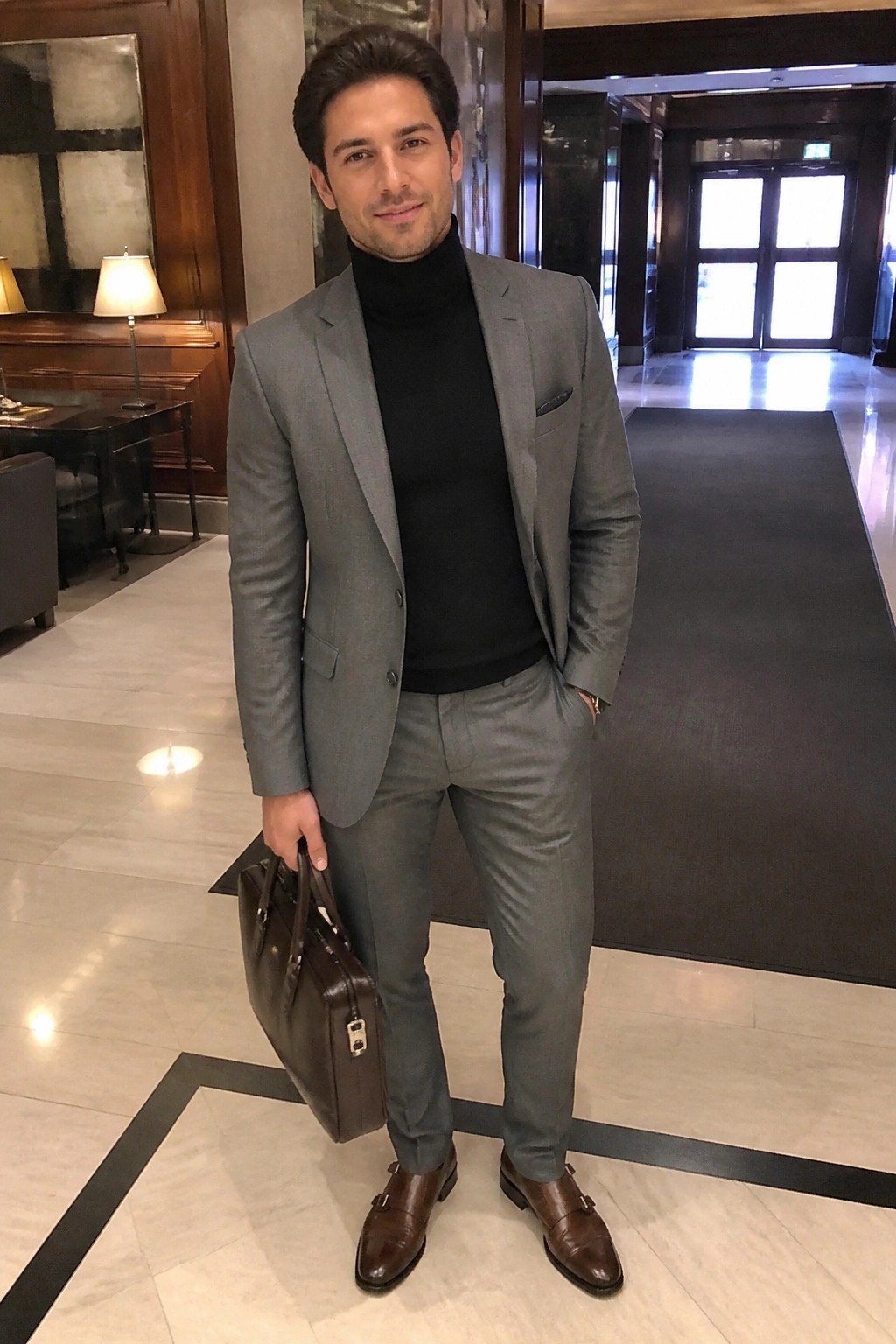 Man in gray suit and black turtleneck holding brown briefcase in marble lobby.