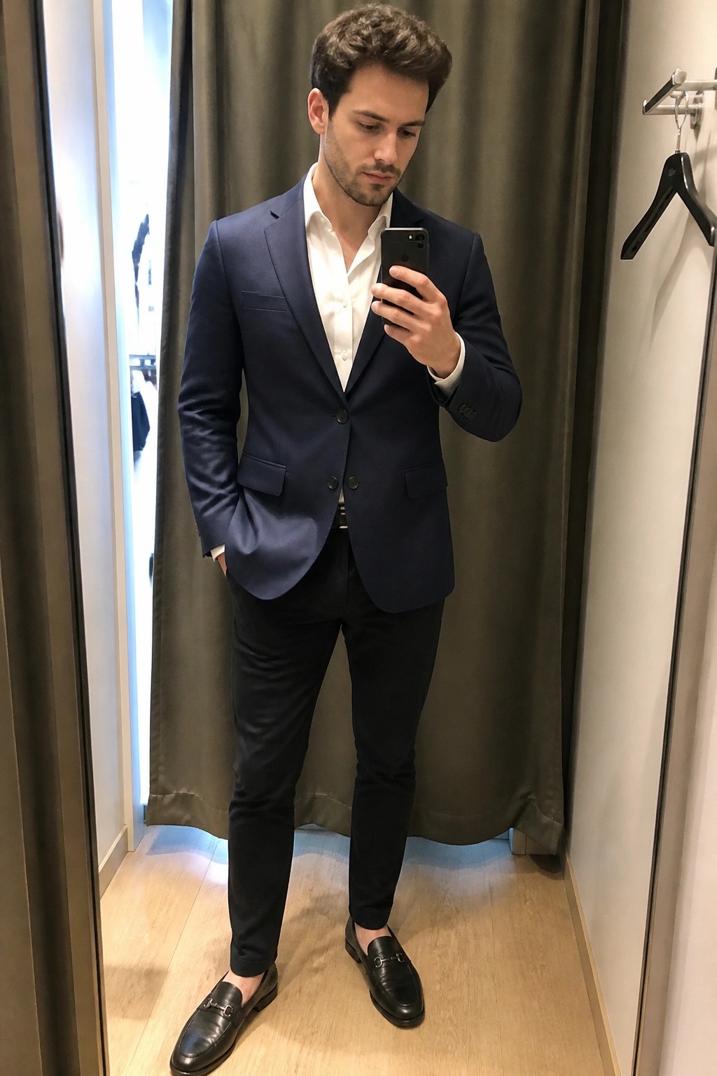 Man in navy blazer and black pants taking mirror selfie in fitting room