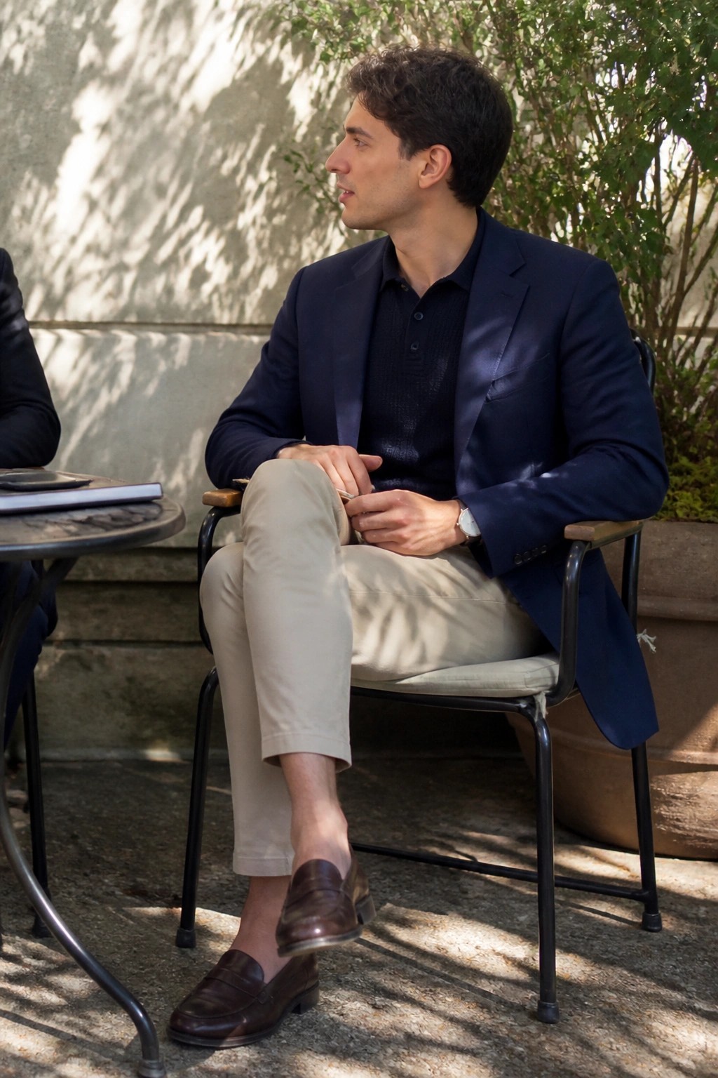 Man in navy blazer and beige pants sits in sunny outdoor chair.
