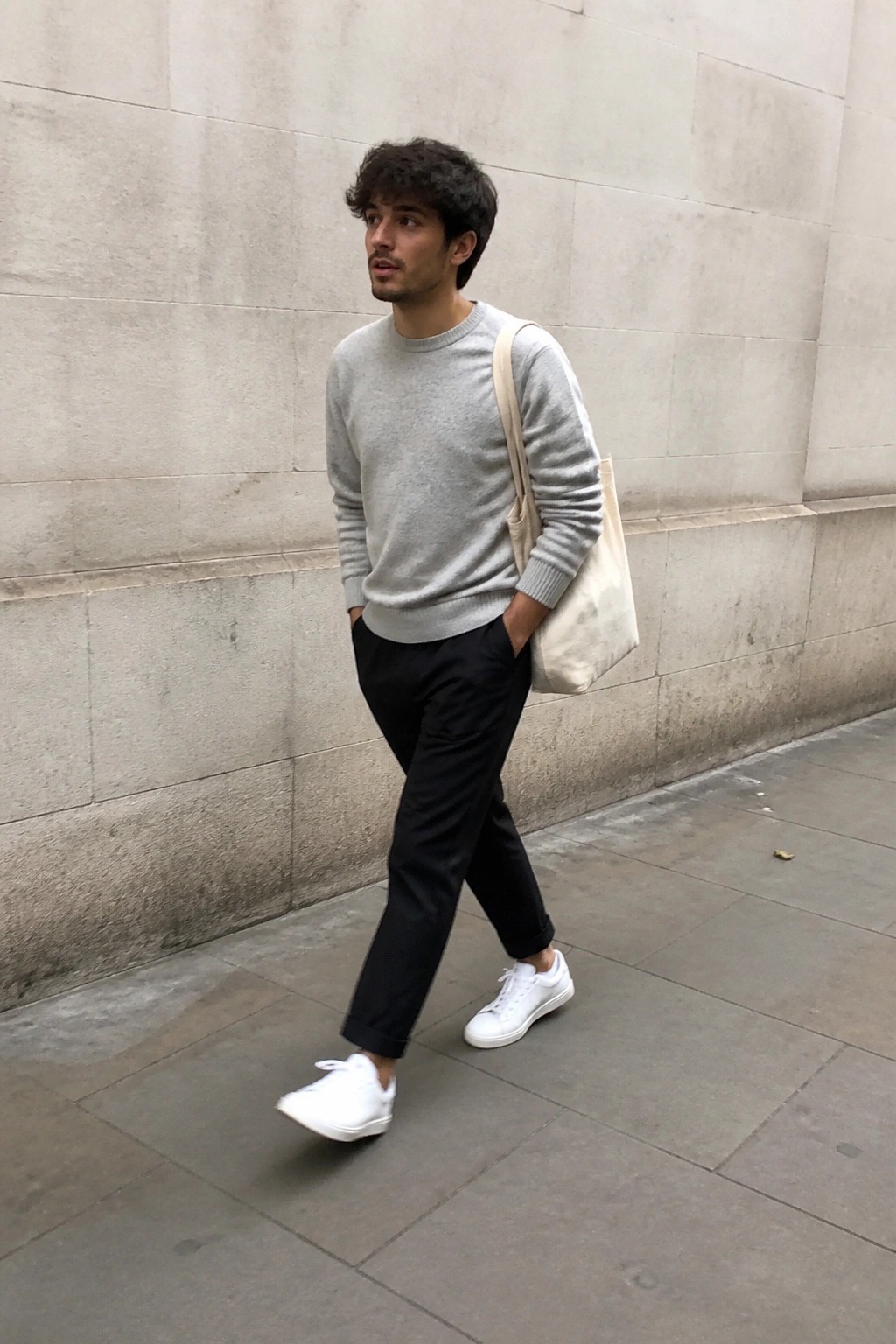 Young man in gray sweater, black pants, white sneakers carries beige tote on sidewalk