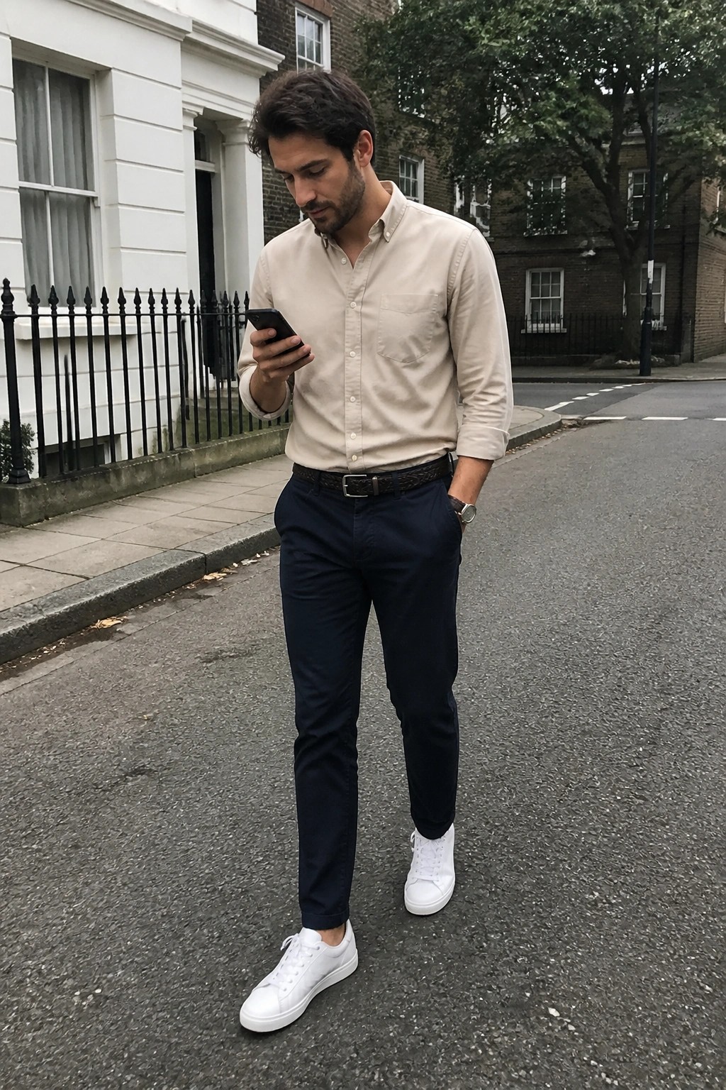 Man in beige shirt and navy pants checks smartphone on urban street