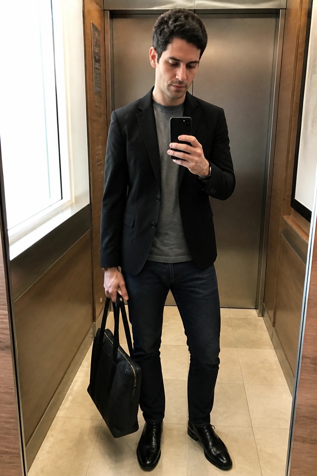 Man in black blazer and jeans holding briefcase and phone in elevator