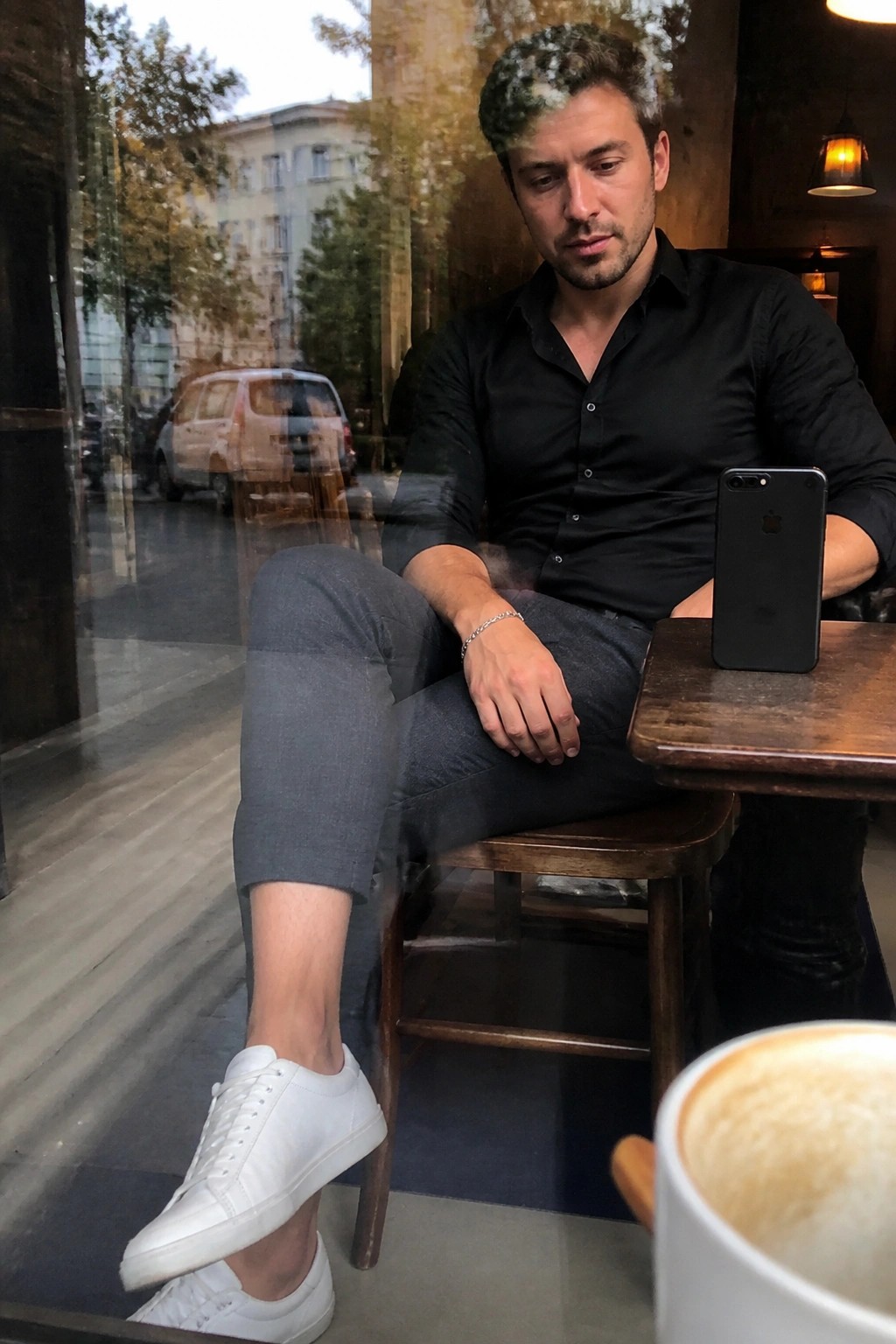 Man in black shirt and grey pants at cafe table with smartphone and coffee