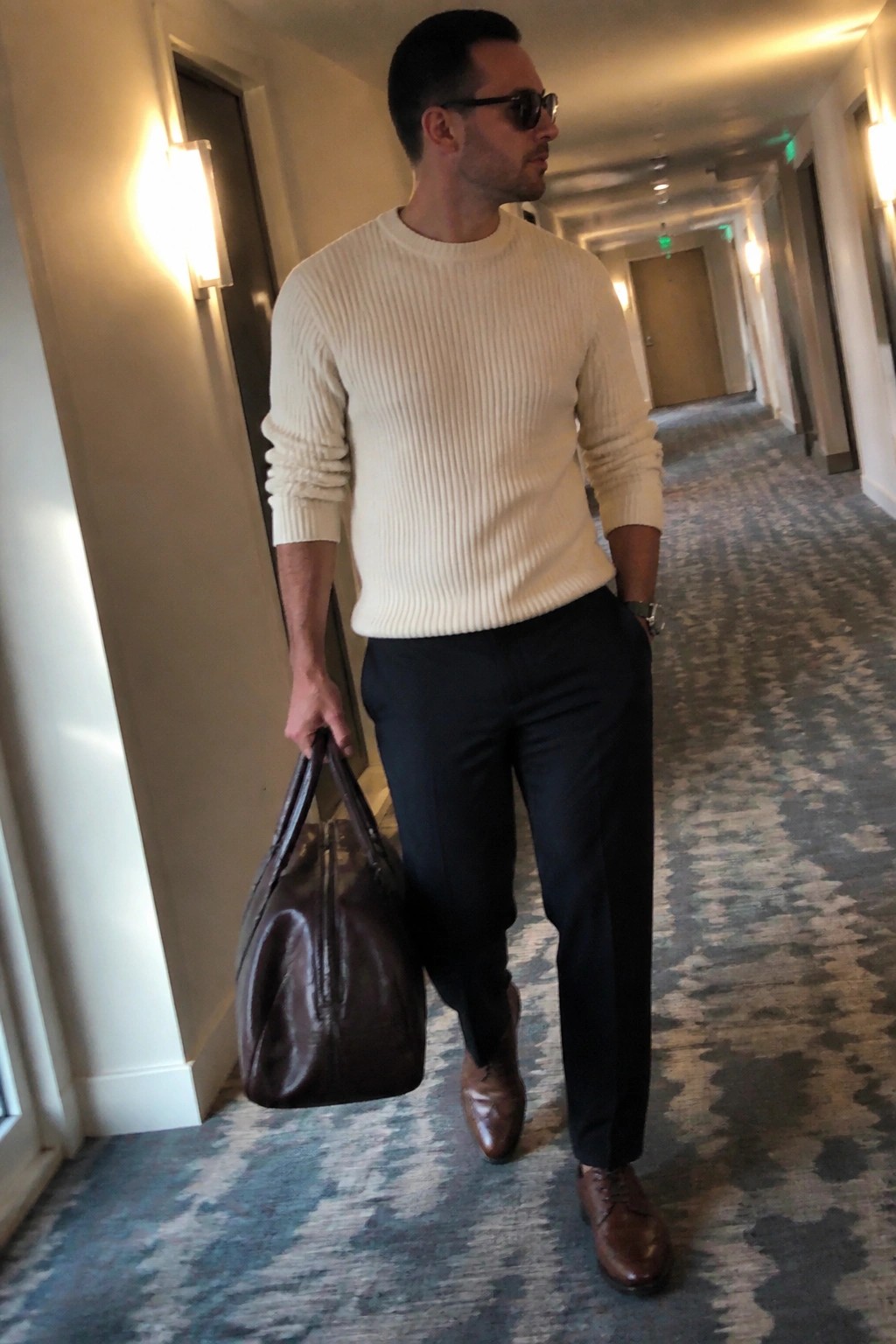 Man in cream ribbed sweater and black pants holds brown leather bag in hallway