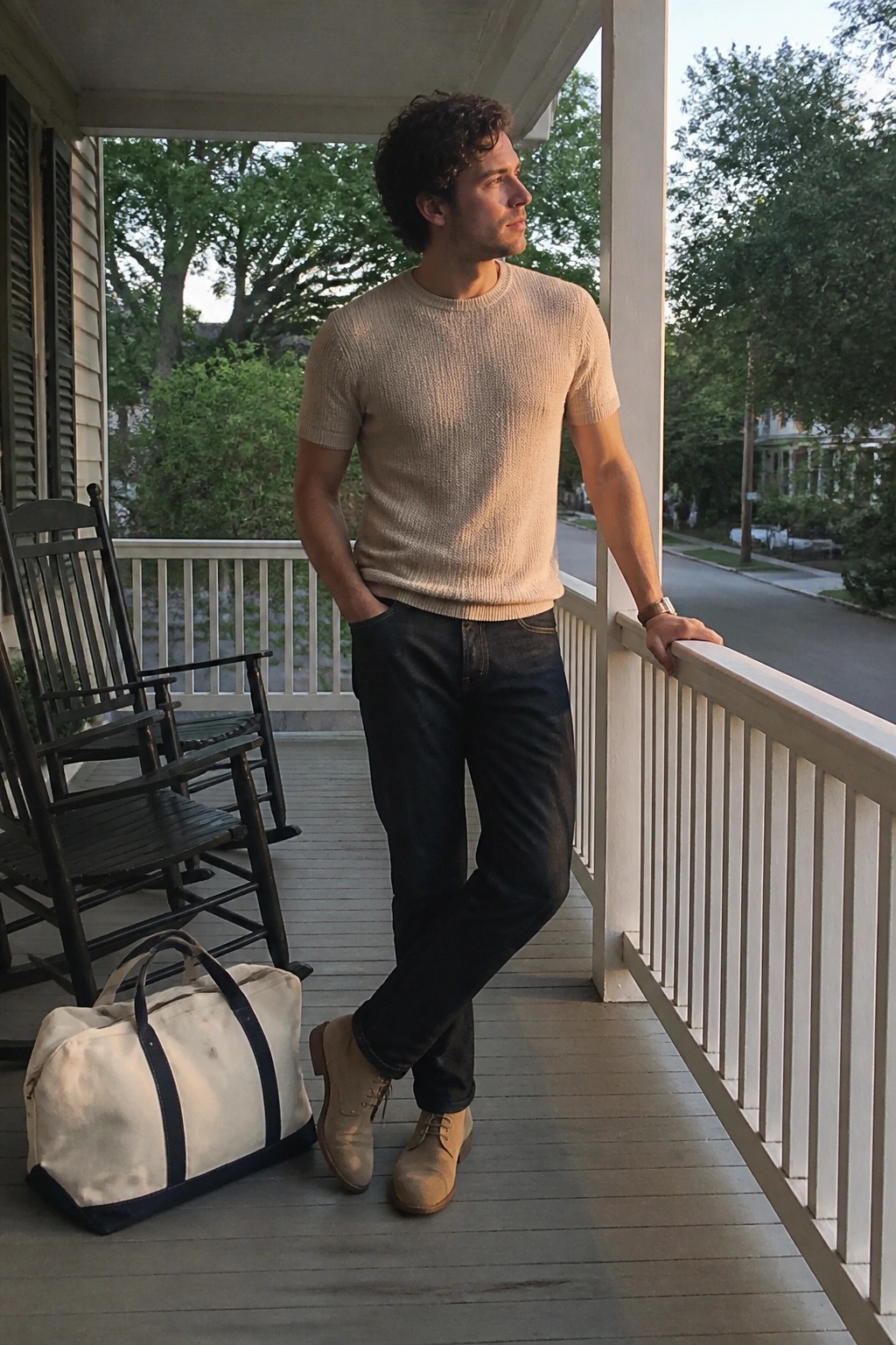 Man in beige sweater leans on porch railing near rocking chair and canvas bag