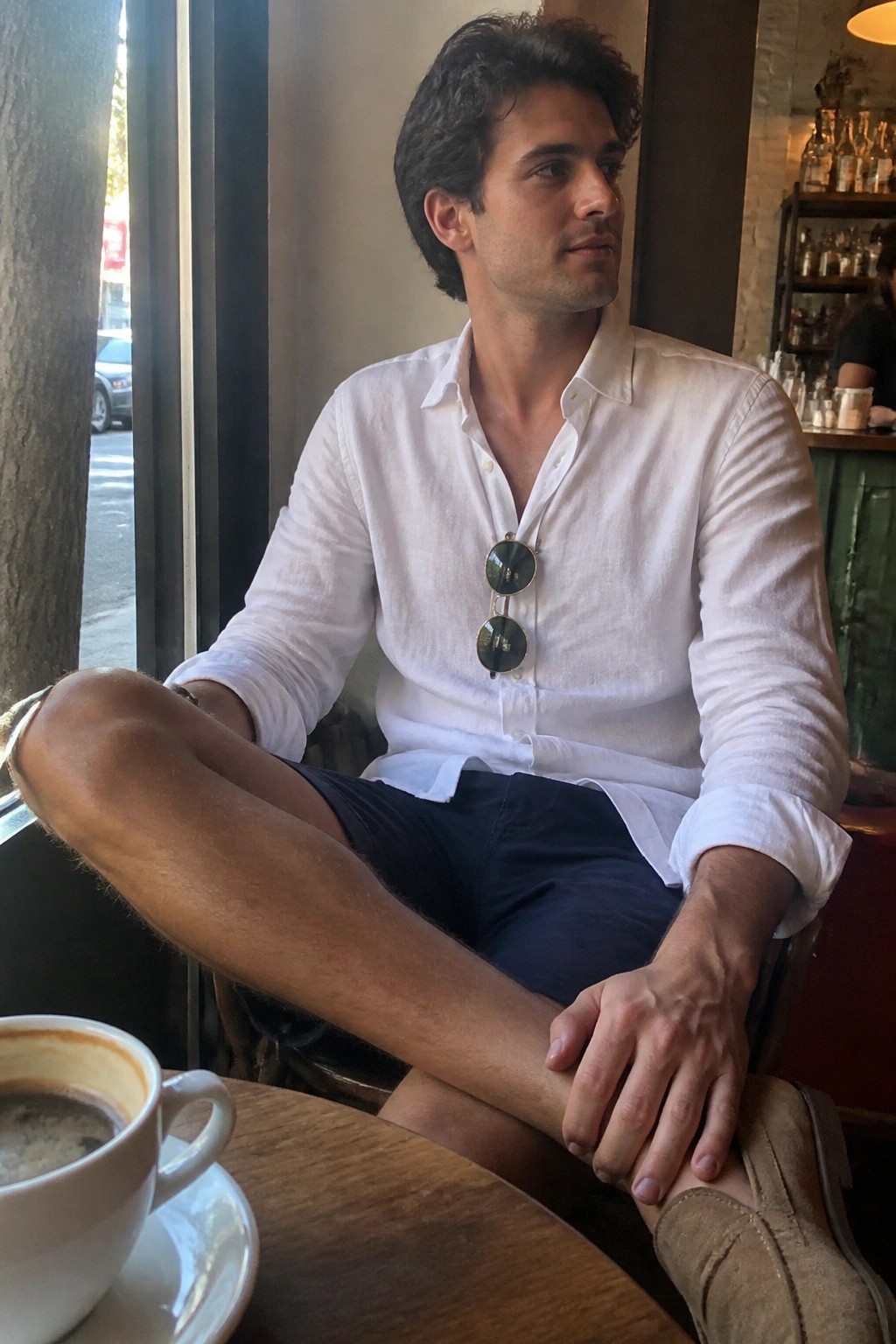 Man in white shirt with sunglasses, navy shorts, seated at cafe table by window