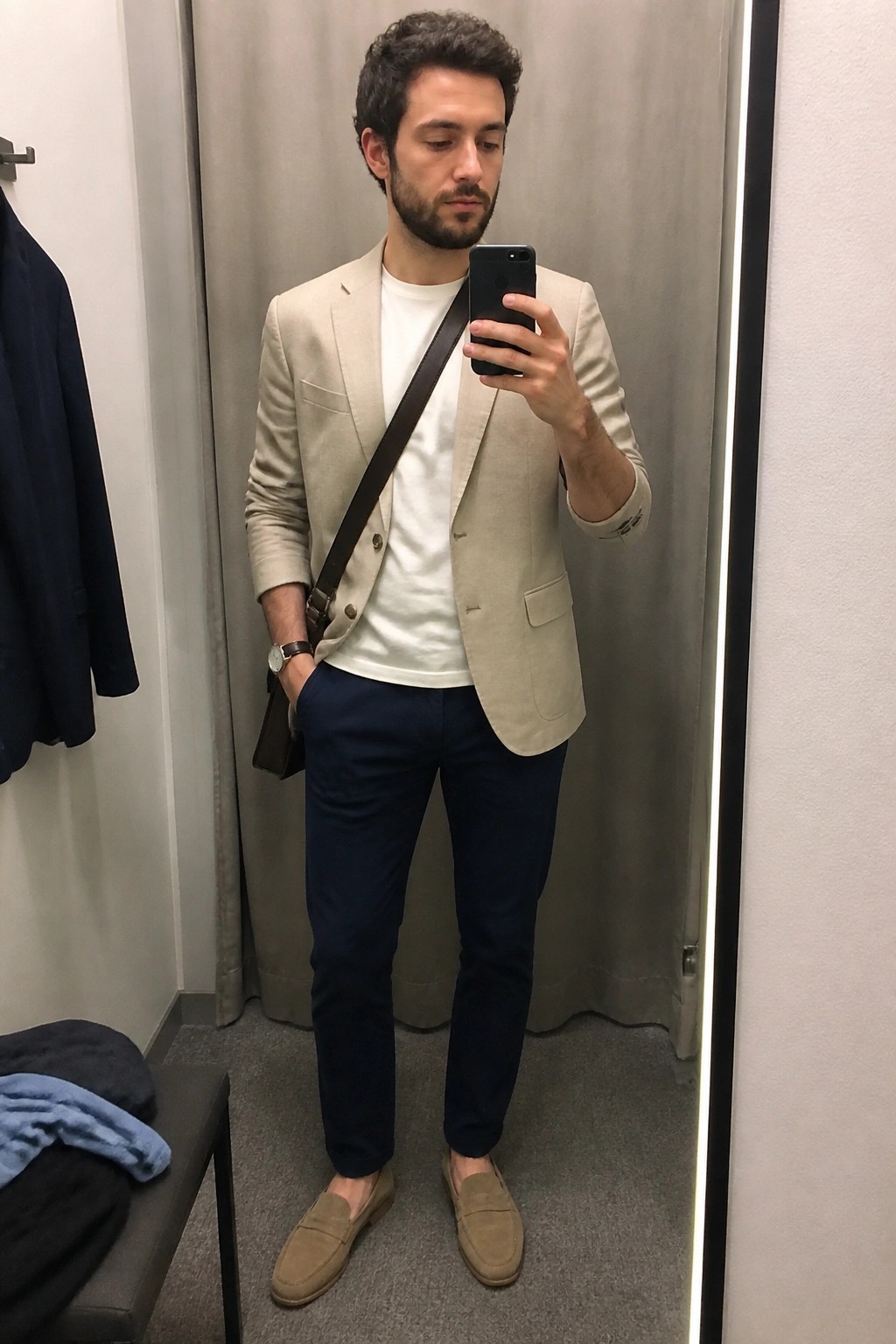 Man in beige blazer over white tee with navy pants takes mirror selfie in fitting room
