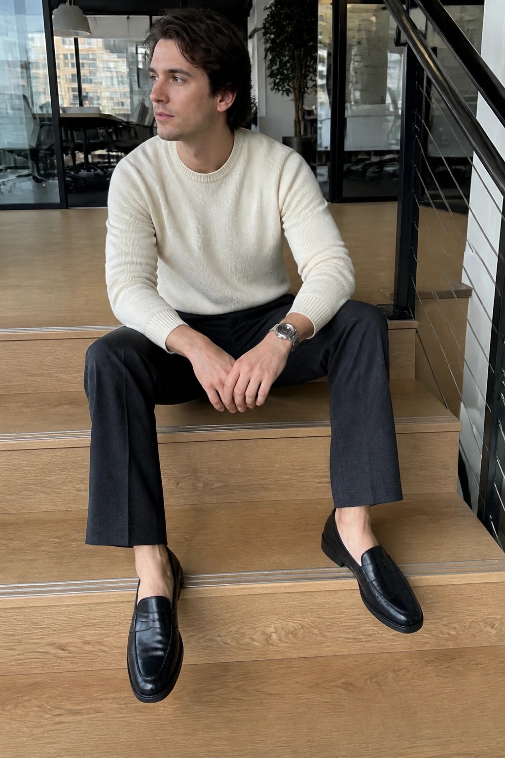 Man in cream sweater and black pants seated on wooden stairs wearing loafers