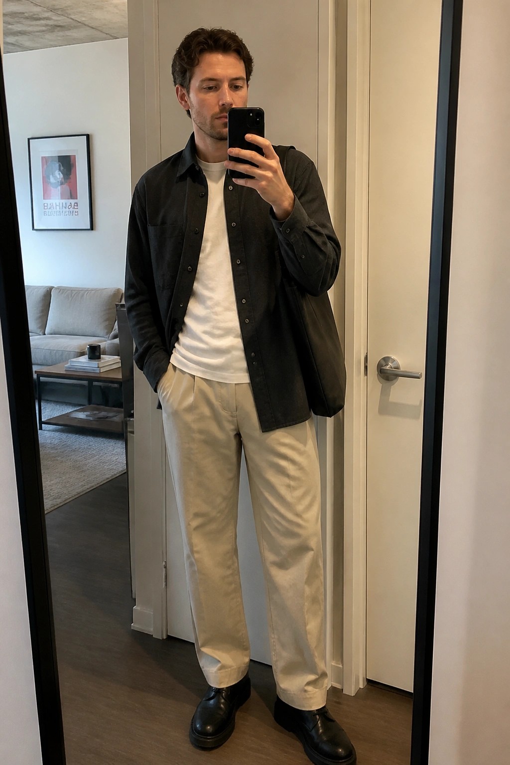 Man in dark button-up over white tee with beige pants taking mirror selfie in apartment