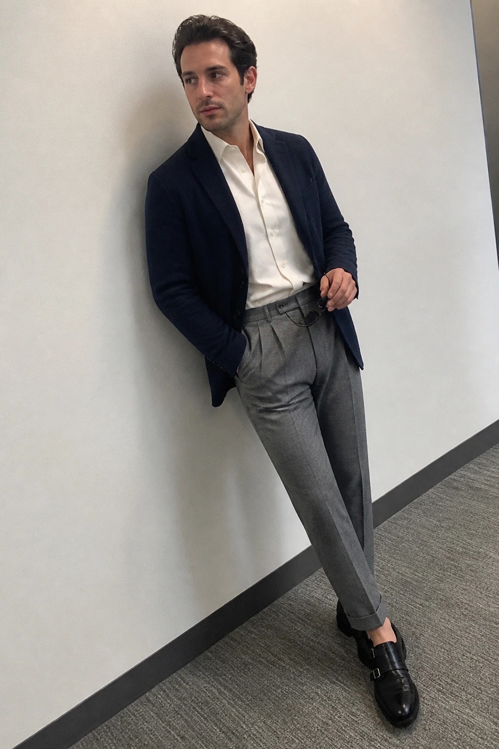 Man in navy blazer, white shirt, gray trousers, and black monk shoes leaning on wall