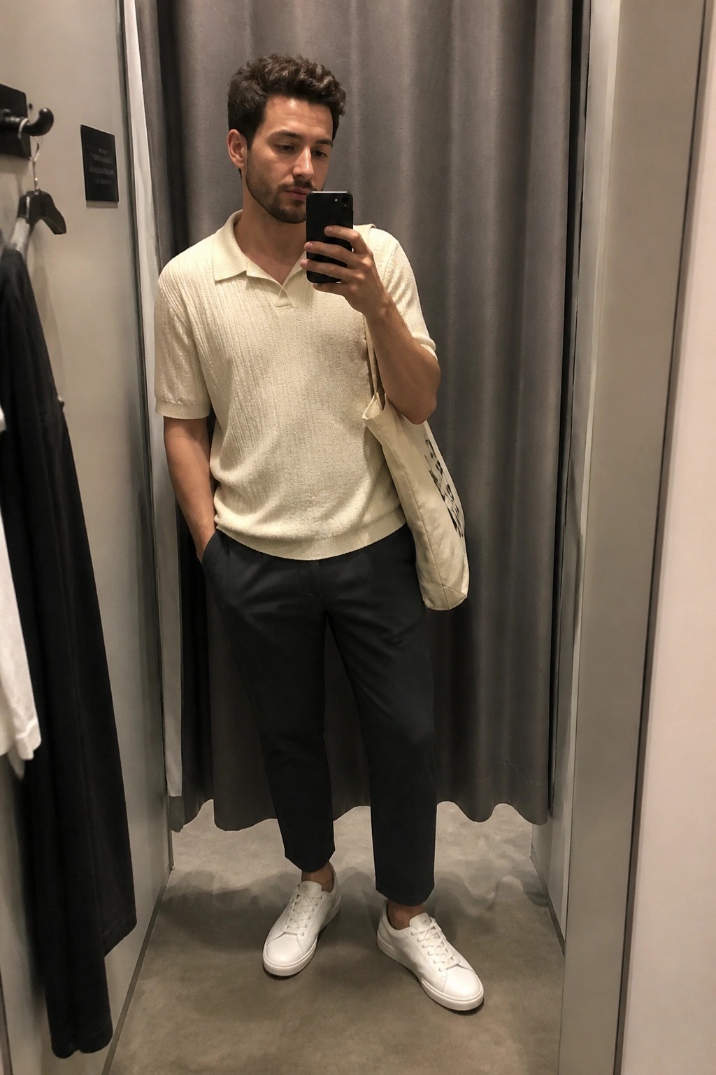 Man in cream polo shirt and dark pants taking selfie in fitting room with tote bag