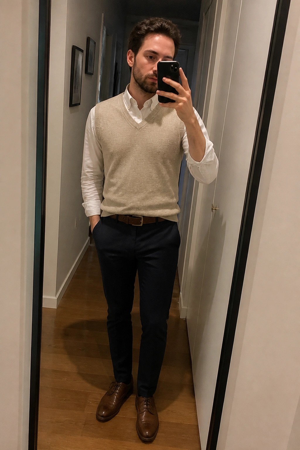 Man in beige sweater vest and navy pants taking hallway mirror selfie with phone