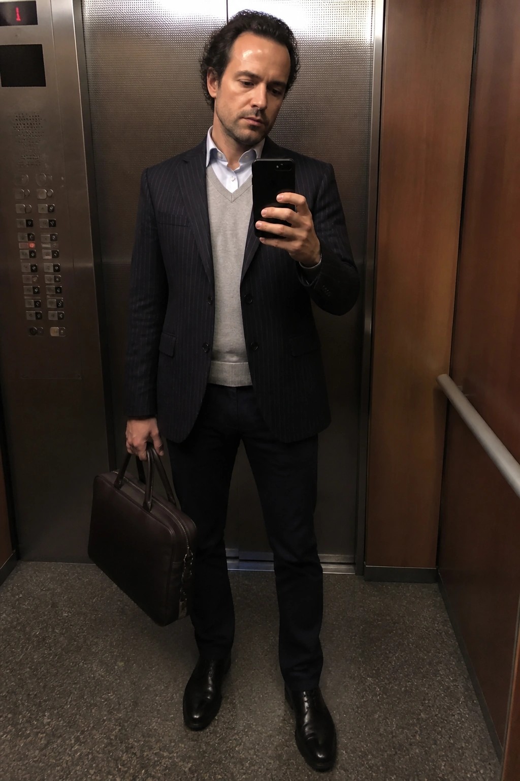 Man in pinstripe suit taking phone selfie in elevator holding leather briefcase.