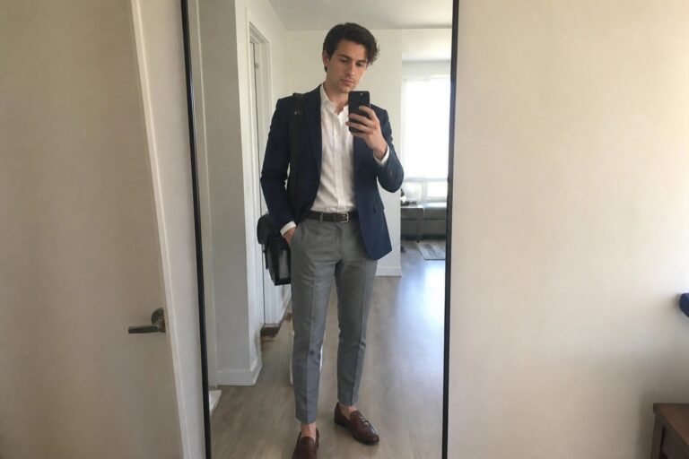 20 Modern Office Outfits for Men Starting Their Professional Careers Man in navy blazer, white shirt, and gray pants taking mirror selfie with phone and bag.