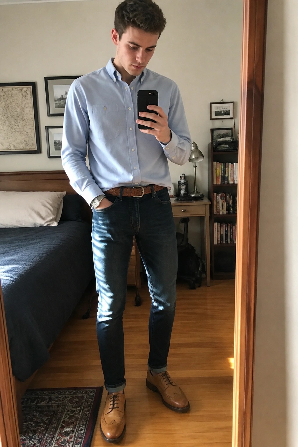 Young man wearing a light blue button down shirt tucked into dark slim jeans with a brown woven belt and brown leather brogue shoes while holding a phone in a bedroom setting.
