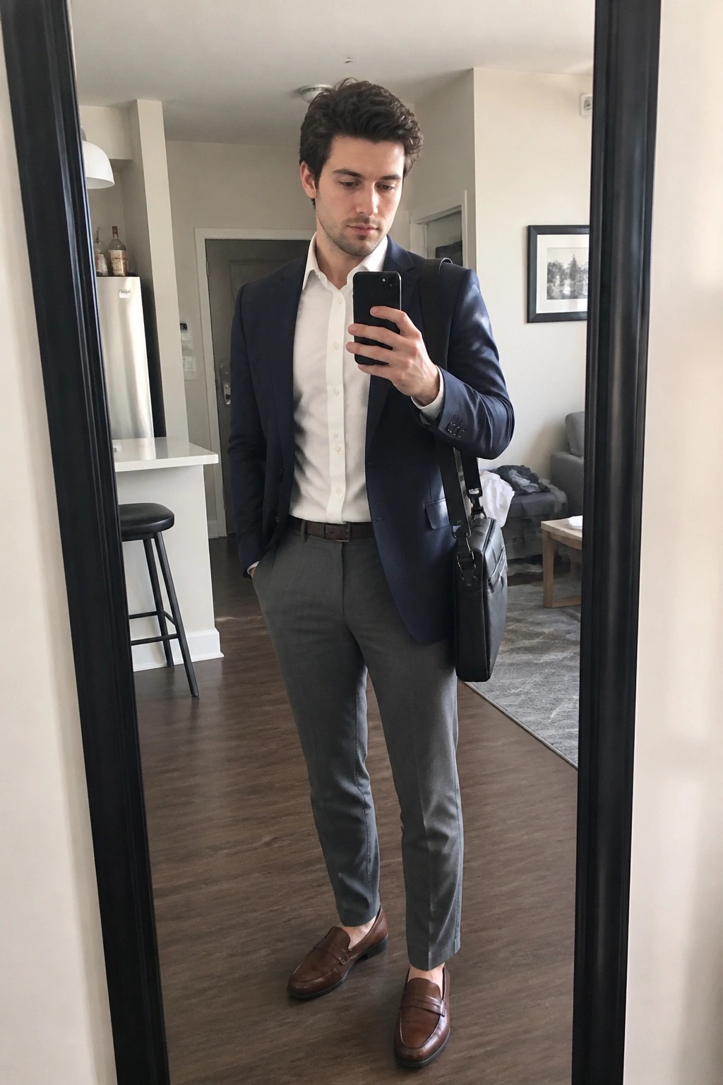 Man in navy blazer, white shirt, and gray pants taking mirror selfie with phone and bag.