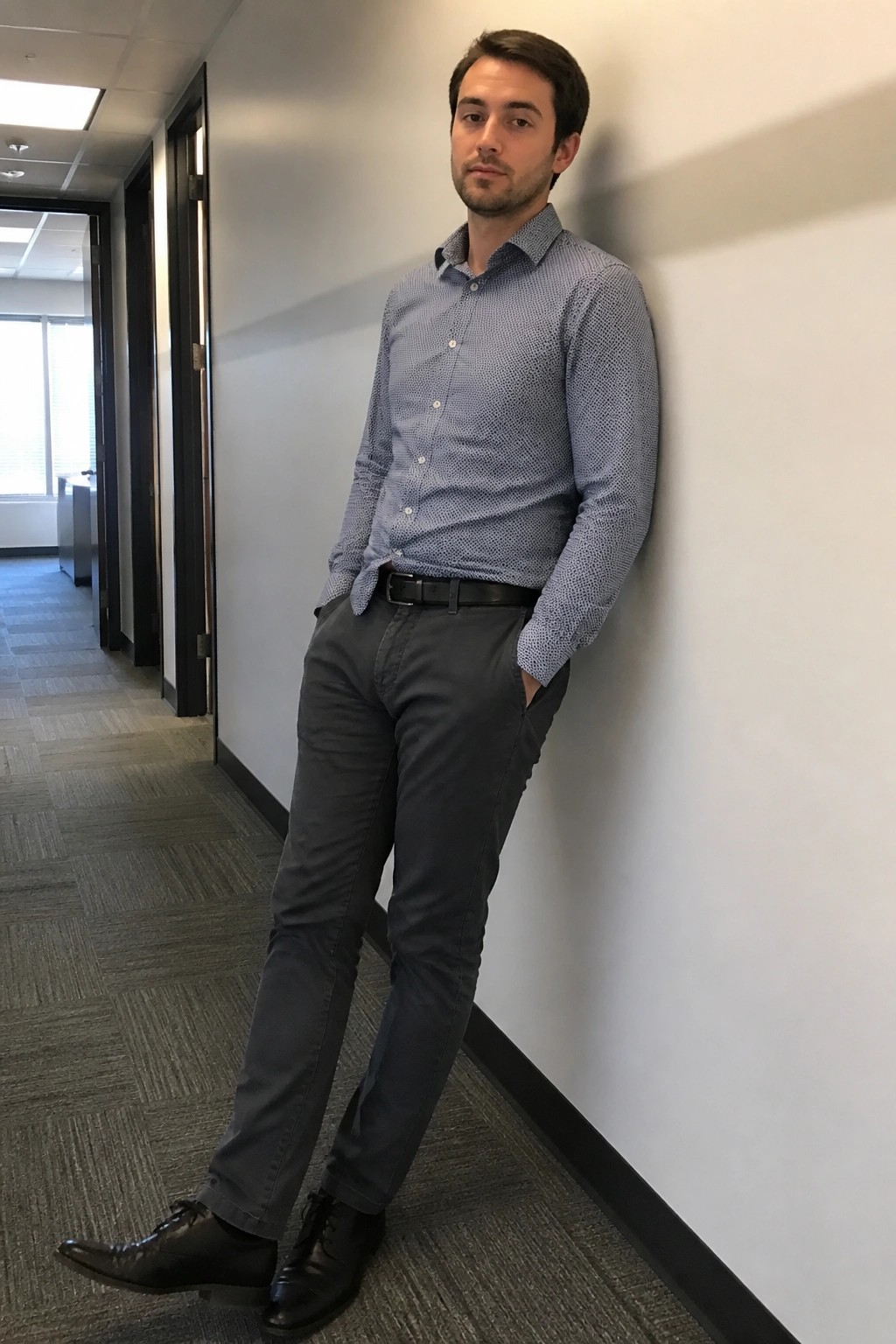 Man in blue patterned shirt and gray pants leaning on office wall