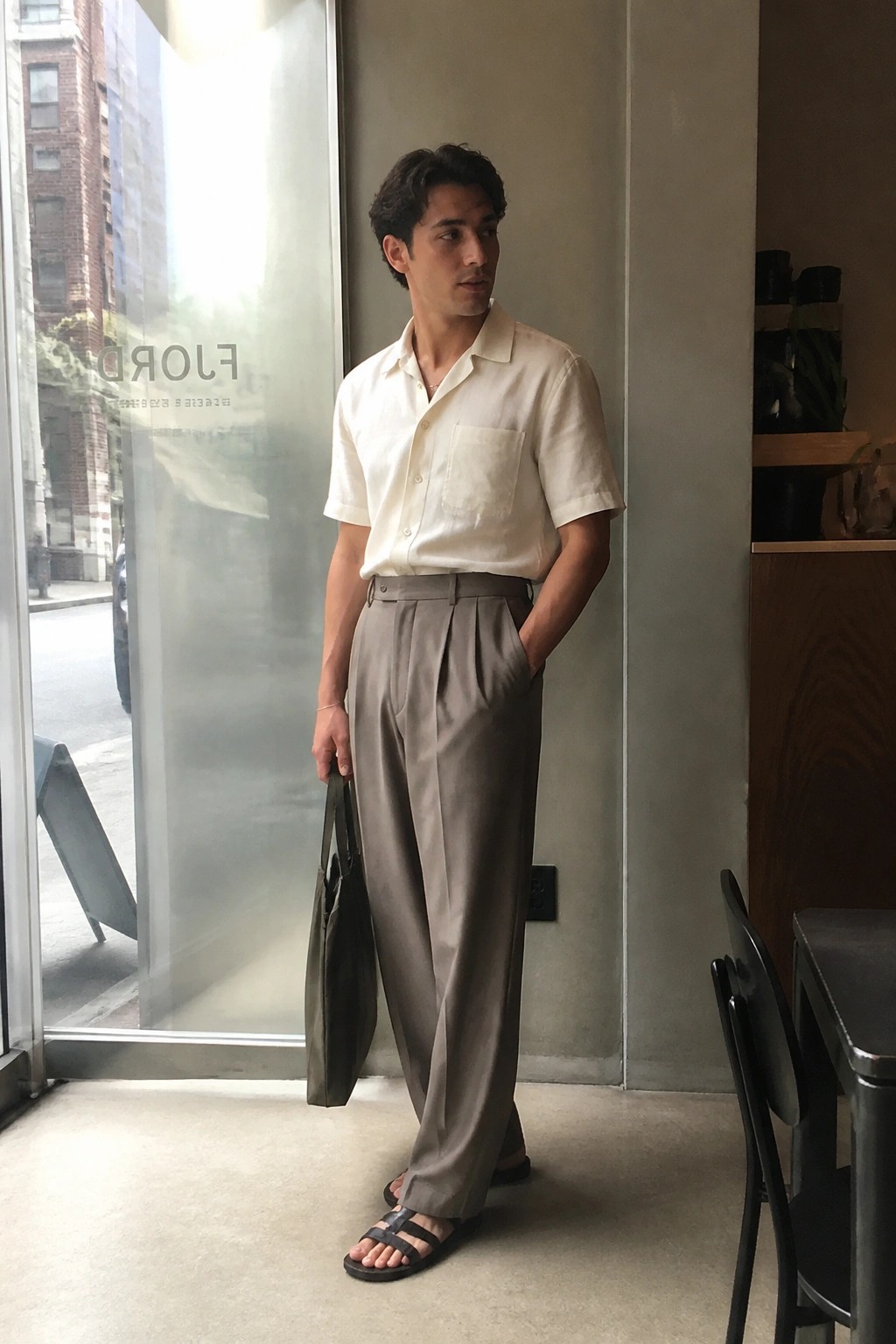 Man in cream short-sleeve shirt and gray pleated trousers holding olive tote bag.
