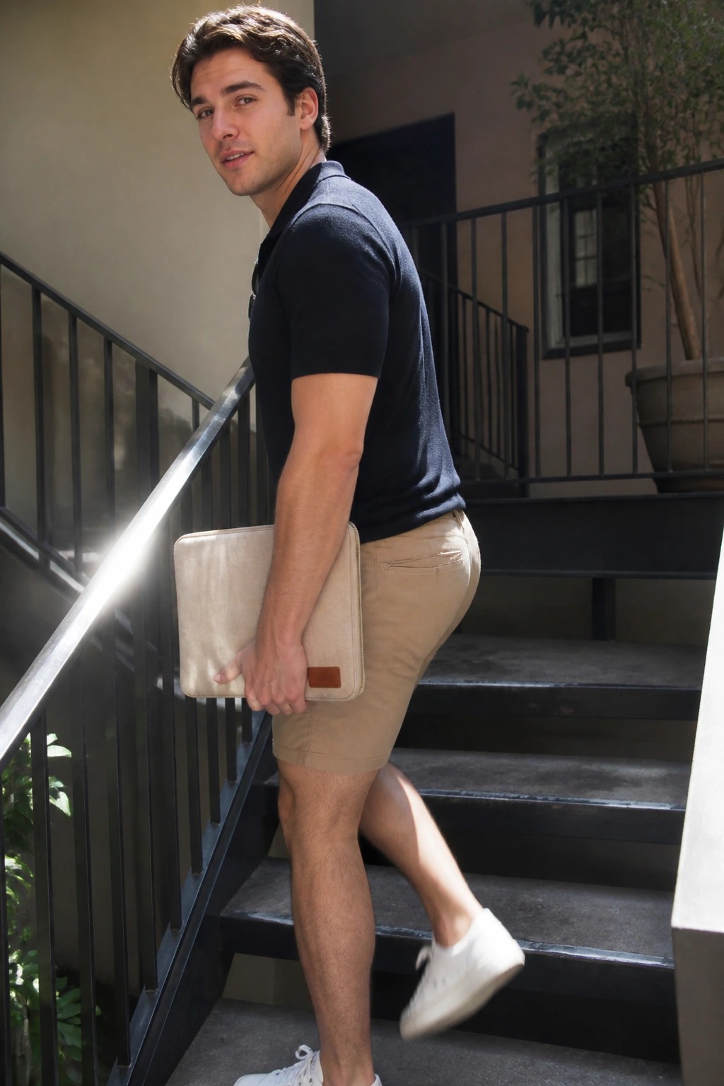 Man in navy polo shirt and khaki shorts holds beige laptop case on stairs