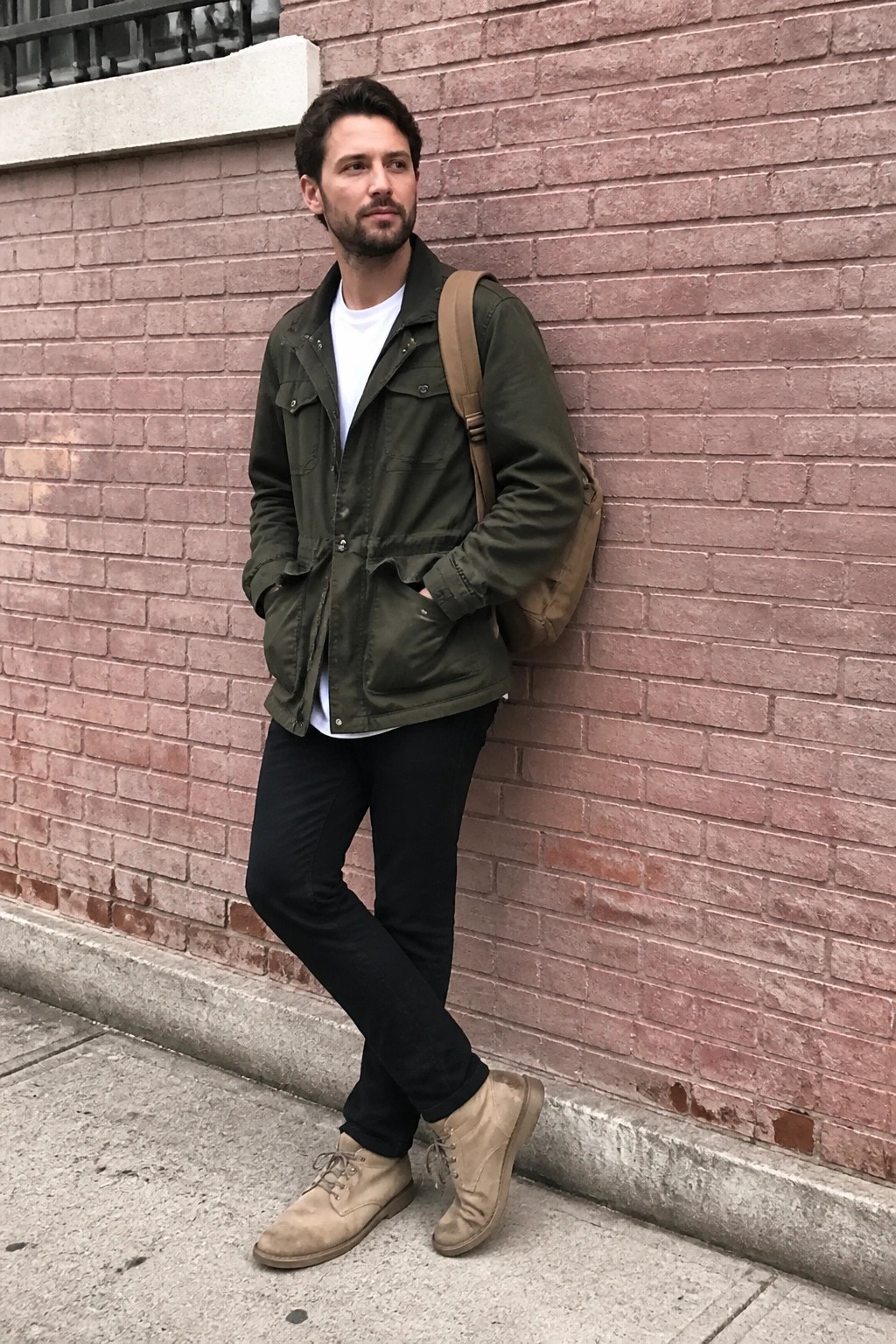 Man in olive jacket and black pants leans against red brick wall with backpack.