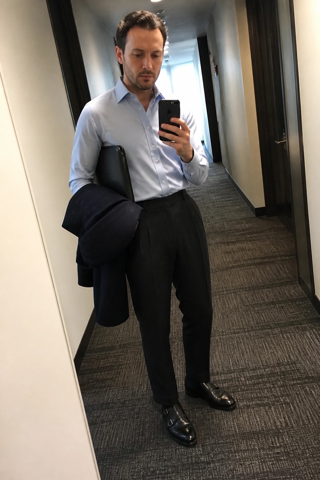 Man in light blue shirt holding phone and jacket in office hallway