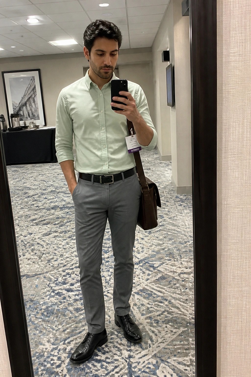 Man in light green shirt and gray pants taking mirror selfie with brown bag