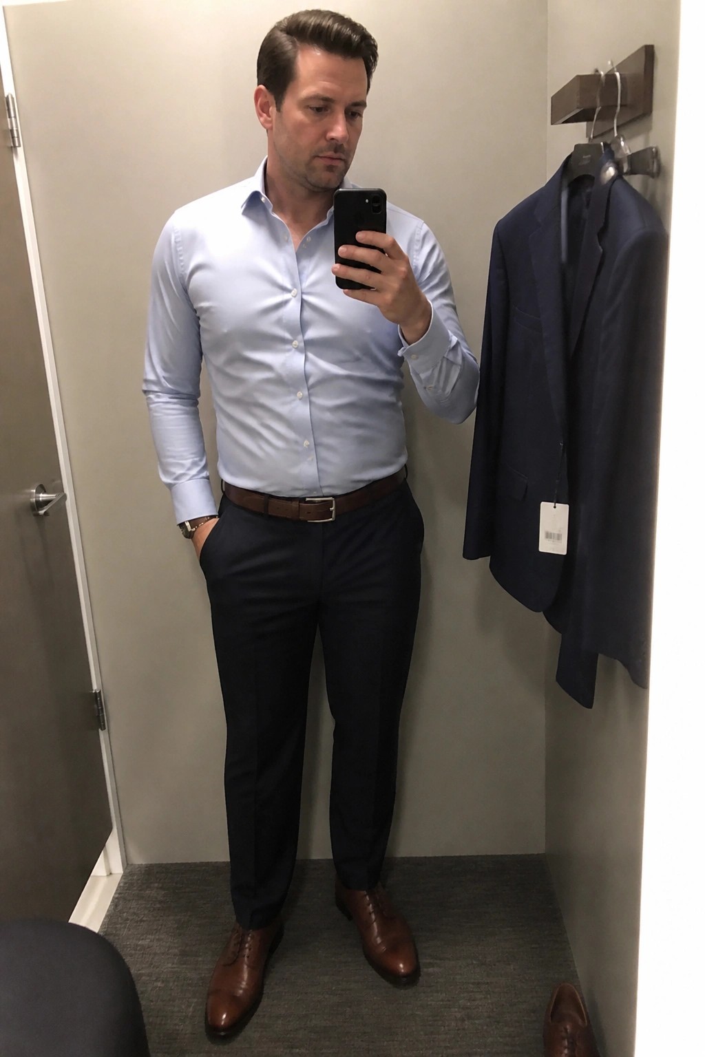 Man wearing light blue button down shirt tucked into navy trousers with brown belt and brown leather shoes while holding a phone in a fitting room mirror selfie.